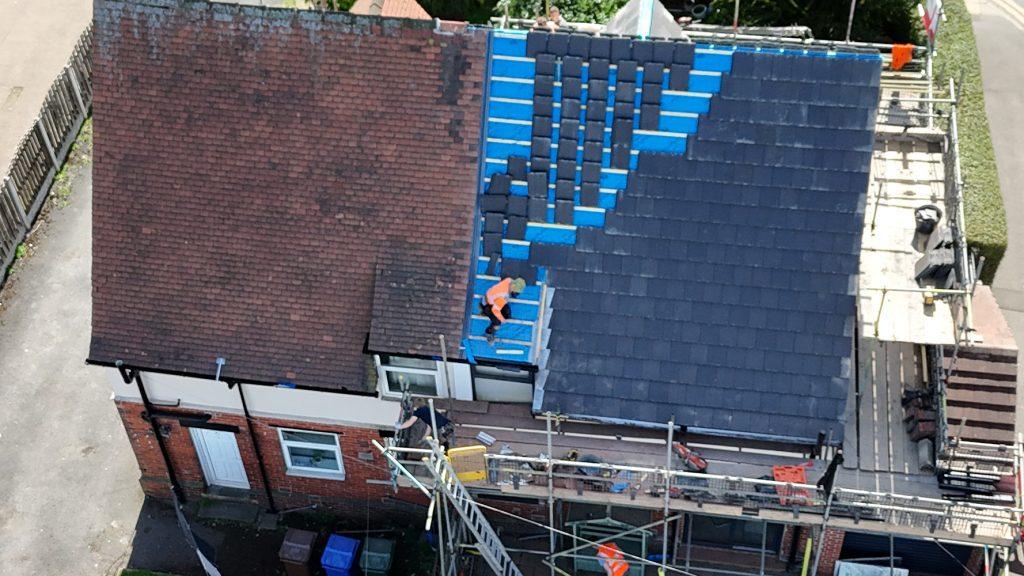 Roofer installing dark roof tiles on a house beside an older red tiled roof